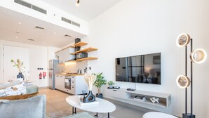 Ein modernes Wohnzimmer mit einer Couch, einem Couchtisch und einem an der Wand befestigten Fernseher. Es gibt eine Küchenzeile mit Holzregalen sowie einen Esstisch mit Stühlen.
