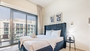 A modern bedroom with a large bed, a small table, and a view of a cityscape through large windows. - Studio Near Meydan Racecourse, Minutes to Downtown, Azizi Riviera 47 by Deluxe Holiday Homes (Dubai)