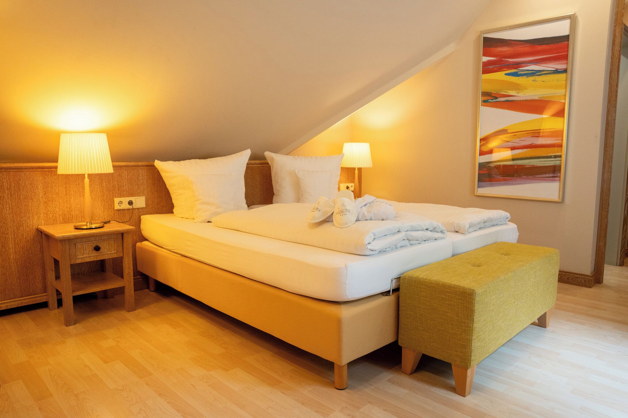 Una habitación de hotel con una cama grande, dos mesitas de noche con lámparas, un banco y un cuadro abstracto y colorido en la pared.