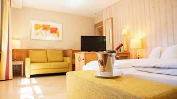 Una habitación de hotel con un sofá amarillo, una cama con ropa blanca, un televisor sobre un mueble de madera y un cuadro en la pared.