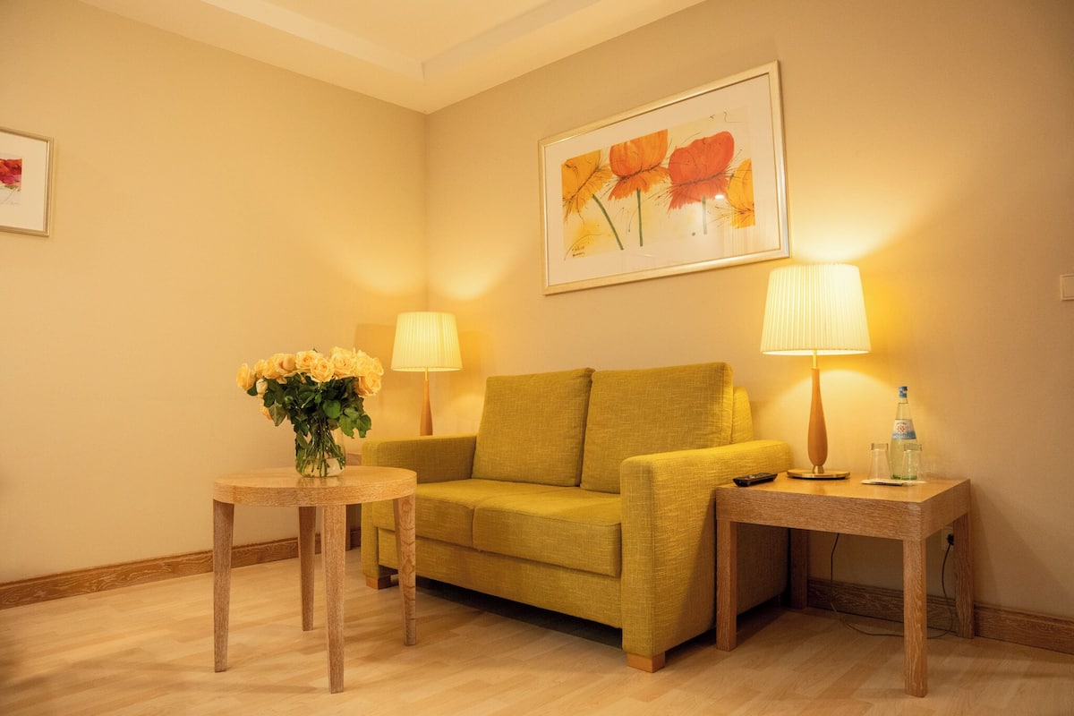 A cozy room with a yellow sofa, two side tables with lamps, a vase of flowers, and a framed floral painting on the wall.