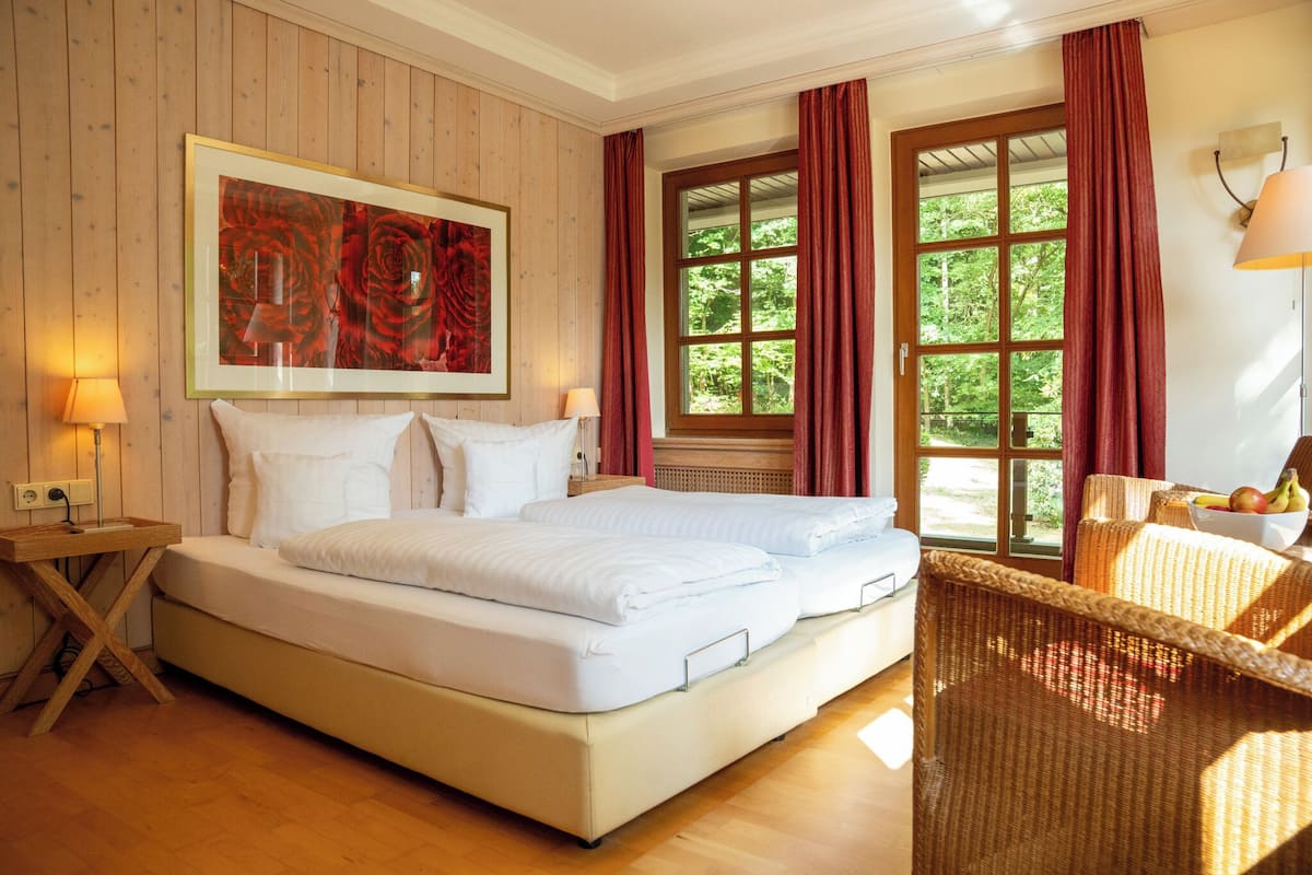 A bedroom with a large bed, wooden walls, a window with curtains, a bedside table with a lamp, and a wicker chair.