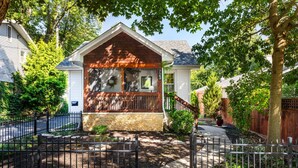 A two-story house with a stone foundation, a wooden front porch, and a black metal fence. - Charming 3BR Cottage + Garden, Walk to Downtown & Metra, Sleeps 6 (Geneva)