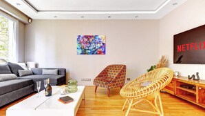 A modern living room with a grey sofa, a coffee table with books and glasses, a colorful abstract painting, a wicker chair, and a wooden TV stand with a Netflix sign. - Appartement chic avec terrasse -  Champs-Elysées (Paris)