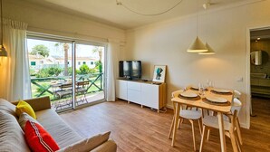 A modern living room with a sofa, a dining table set for two, a TV, and a balcony with a view of houses and greenery. - Sun Lovers Altura T1 Pool & Balcony (Altura)