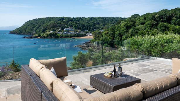 Una terraza con mobiliario de mimbre, una mesa de vidrio y una vista panorámica del mar y una colina boscosa.