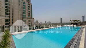 A swimming pool with a clear blue water surface, surrounded by a stone deck and a row of lounge chairs. In the background, there are high-rise buildings and a clear sky. - Bloomfields Cozy 1BR Binghatti Nova  (Dubai)