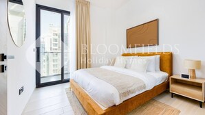 A modern bedroom with a large bed, a nightstand, a lamp, and a view of buildings through a large window. - Bloomfields Cozy 1BR Binghatti Nova  (Dubai)