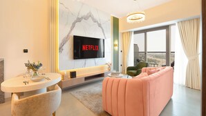 A modern living room with a large flat-screen TV, a pink sofa, a dining table with chairs, and a view of the outdoors through large windows. - KeyRock Living - Golf Gate Apartment  (Dubai)