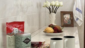 Une salle de bain avec un mur recouvert de carreaux blancs, un vase avec des fleurs blanches, un tableau encadré, une assiette de fruits, une boîte de thon Montego, un sac rouge et deux tasses blanches.