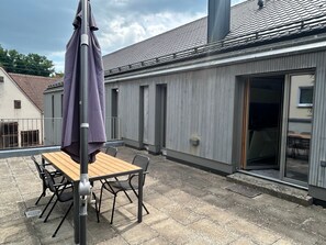 A patio with a table and chairs, a purple umbrella, and a building with a tiled roof and sliding glass doors.