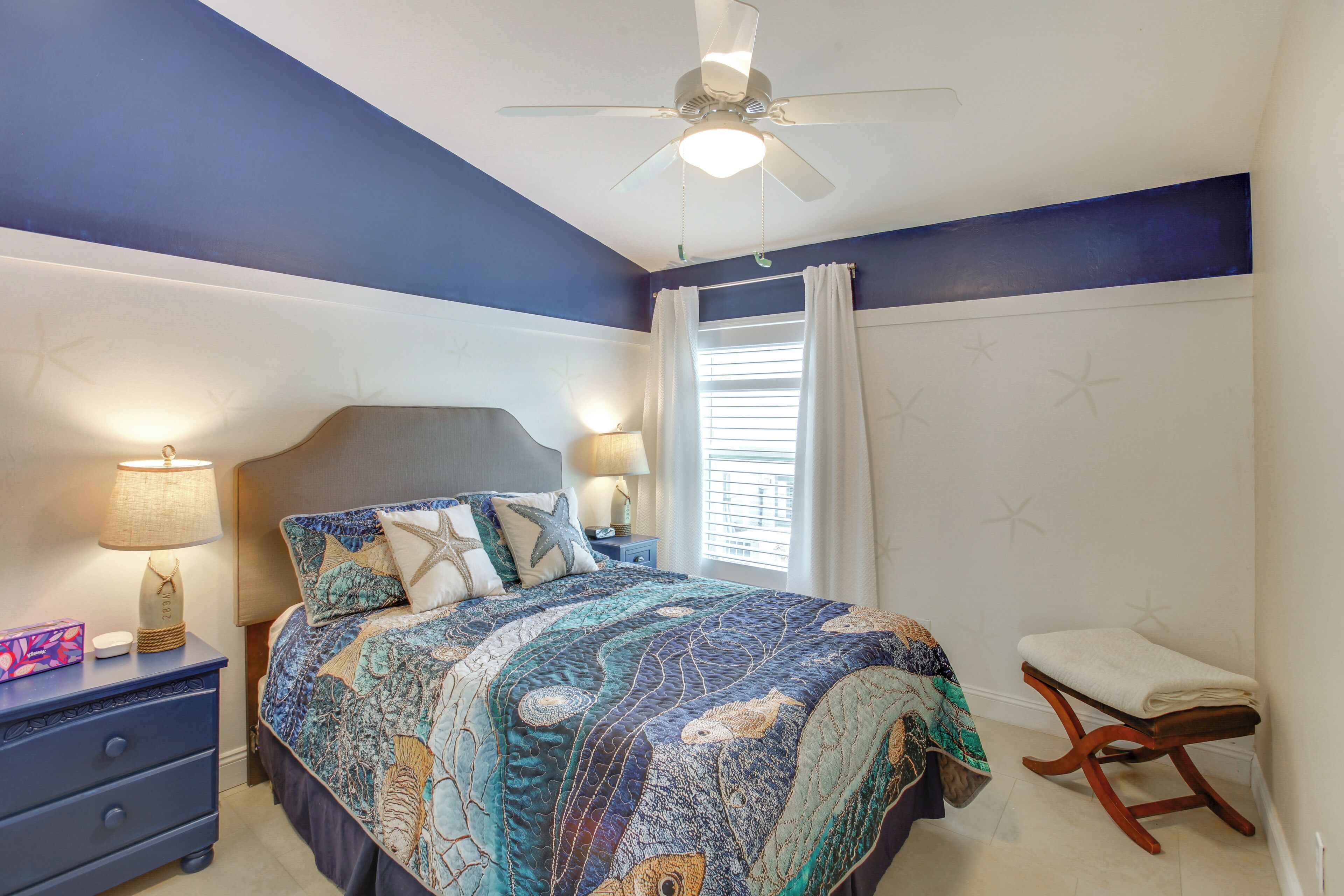 Un dormitorio con cama, mesitas de noche, mesilla de noche, ventilador de techo y ventana con cortinas.