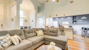 A modern living room with a sectional sofa, a round coffee table with a vase, and a kitchen area with grey cabinets and a bar stool.