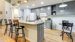 A kitchen with grey cabinets, a white refrigerator, a microwave, and a sink. There are three bar stools and a chair with a frog design.