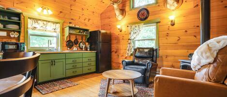 A cozy cabin interior with wooden walls, a dining area with a table and chairs, a kitchen with green cabinets, and a fireplace with a deer head mounted above it.