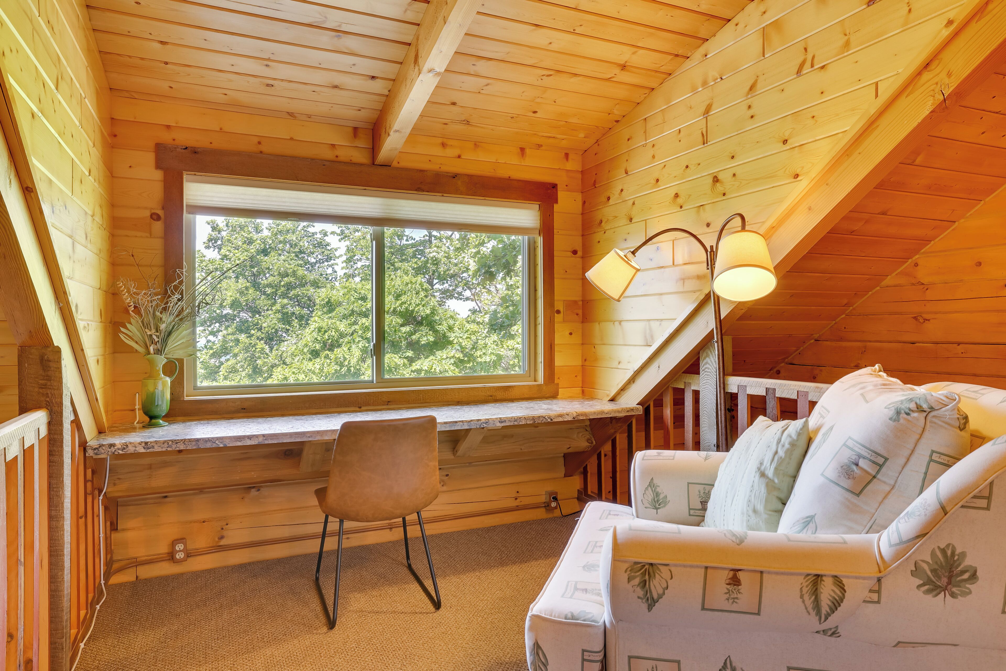 L’intérieur chaleureux d’un chalet avec un plafond en bois, une grande fenêtre, un bureau, une chaise et un coin salon confortable avec des coussins.