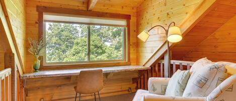 L’intérieur chaleureux d’un chalet avec un plafond en bois, une grande fenêtre, un bureau, une chaise et un coin salon confortable avec des coussins.