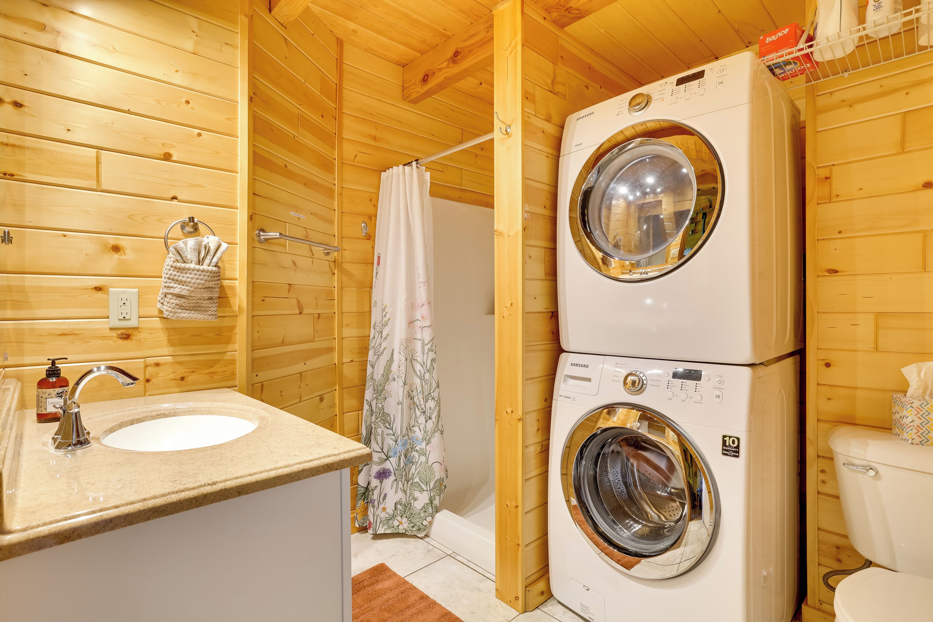 A compact wooden cabin bathroom with a sink, two stacked washing machines, a shower curtain, and a toilet.