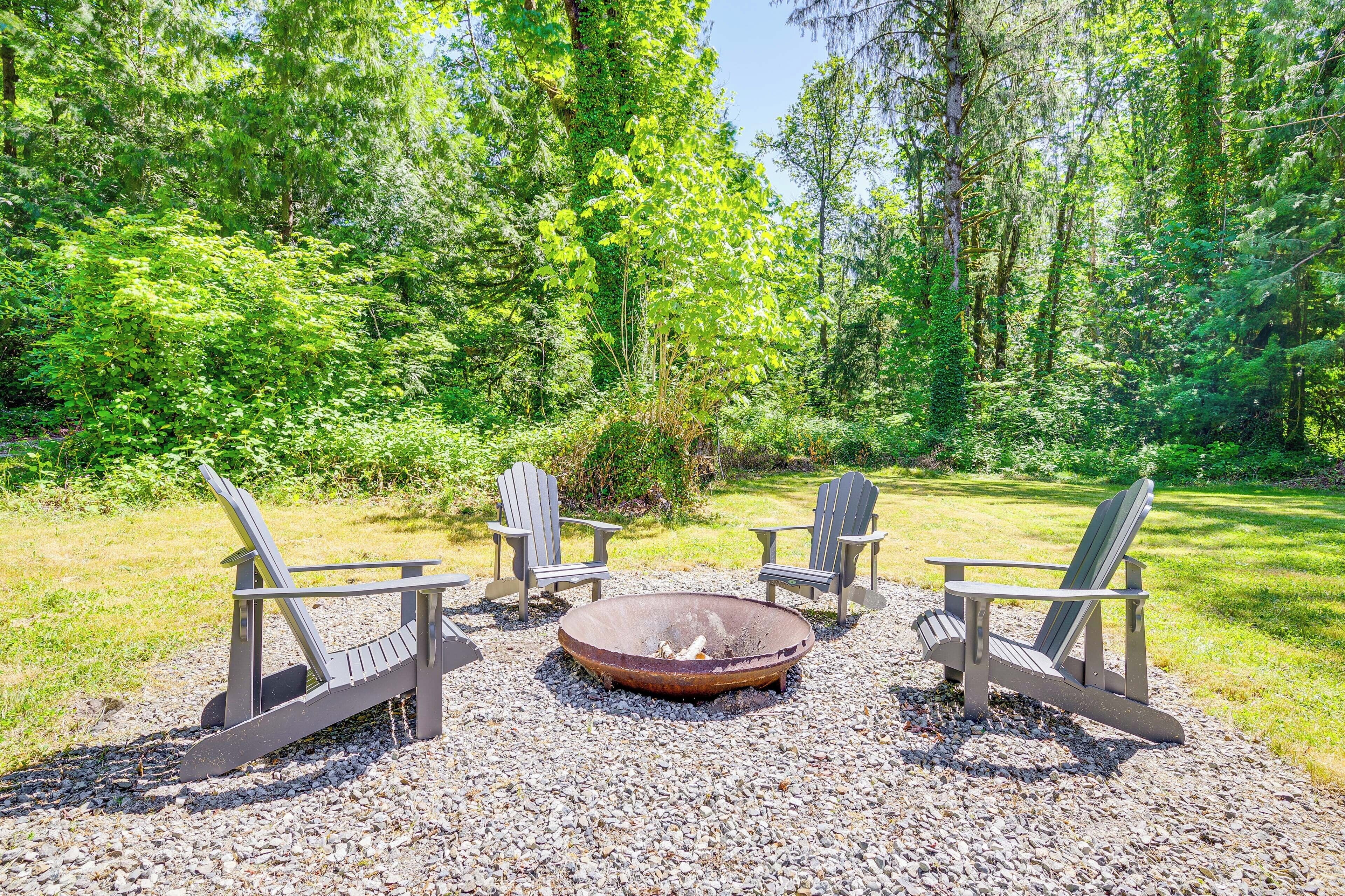 A circular fire pit surrounded by Adirondack chairs in a forest setting.