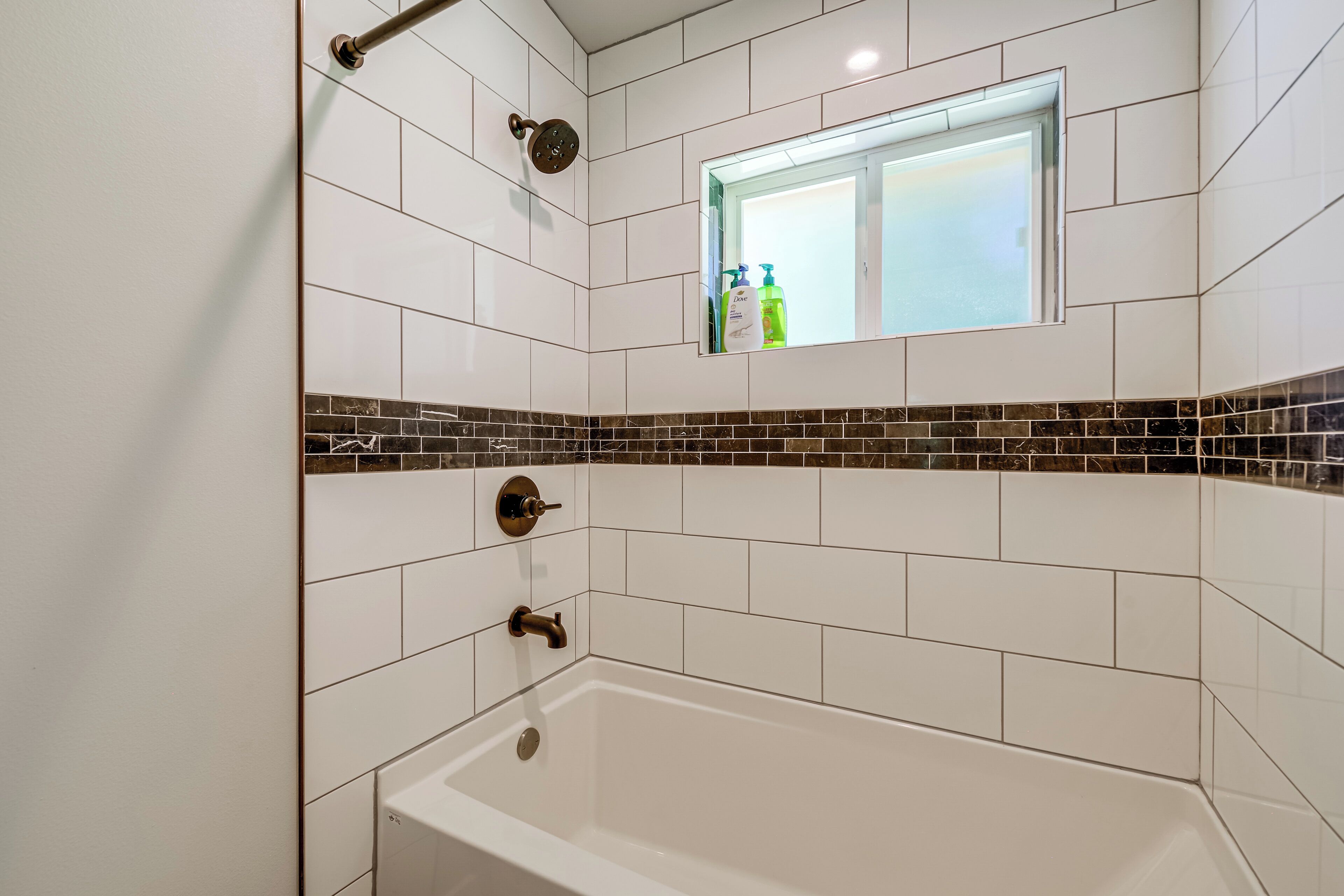A bathroom with white tiles, a shower, a bathtub, and a small window with toiletries.