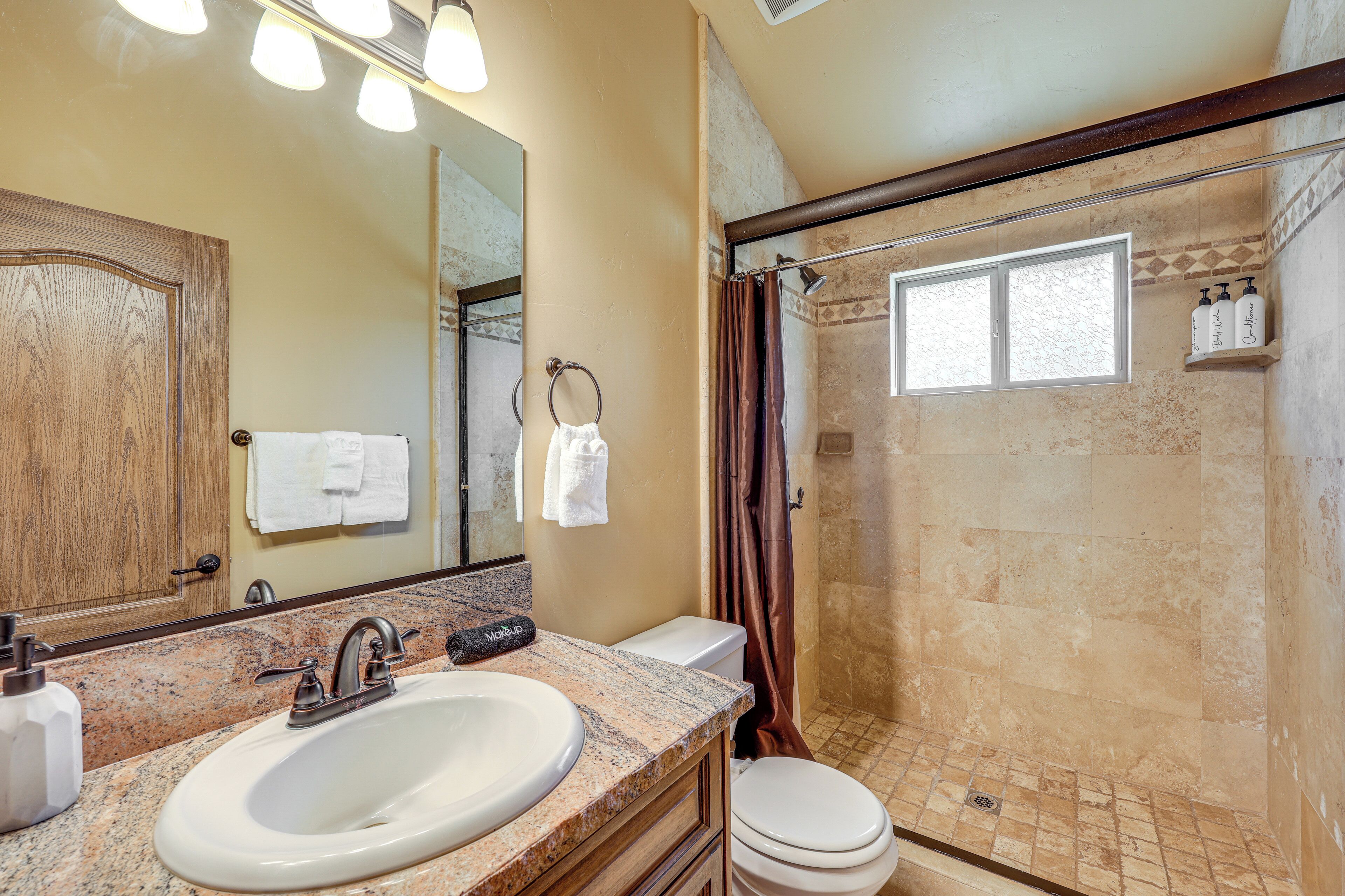 Une salle de bain avec un lavabo sur un plan de travail en granit, un meuble en bois, des toilettes et un espace douche avec une fenêtre.