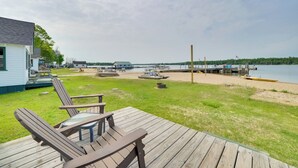 Una terraza de madera con sillas Adirondack, un muelle que se adentra en un lago y un edificio pequeño al fondo.