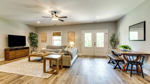 A spacious living room with hardwood flooring, a ceiling fan, a flat-screen TV, a sofa, a coffee table, a dining table with chairs, and two potted plants.