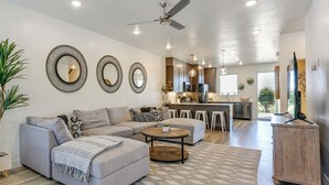 Una sala de estar moderna con un sofá gris, una mesa de centro y un comedor con taburetes de bar. Hay un ventilador de techo, una cocina al fondo y un ventanal que proporciona luz natural.