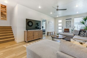 Una sala moderna con un sofá, una mesa de centro y un televisor sobre un mueble de madera. Se ve una escalera que lleva a otra habitación, y un ventilador de techo.