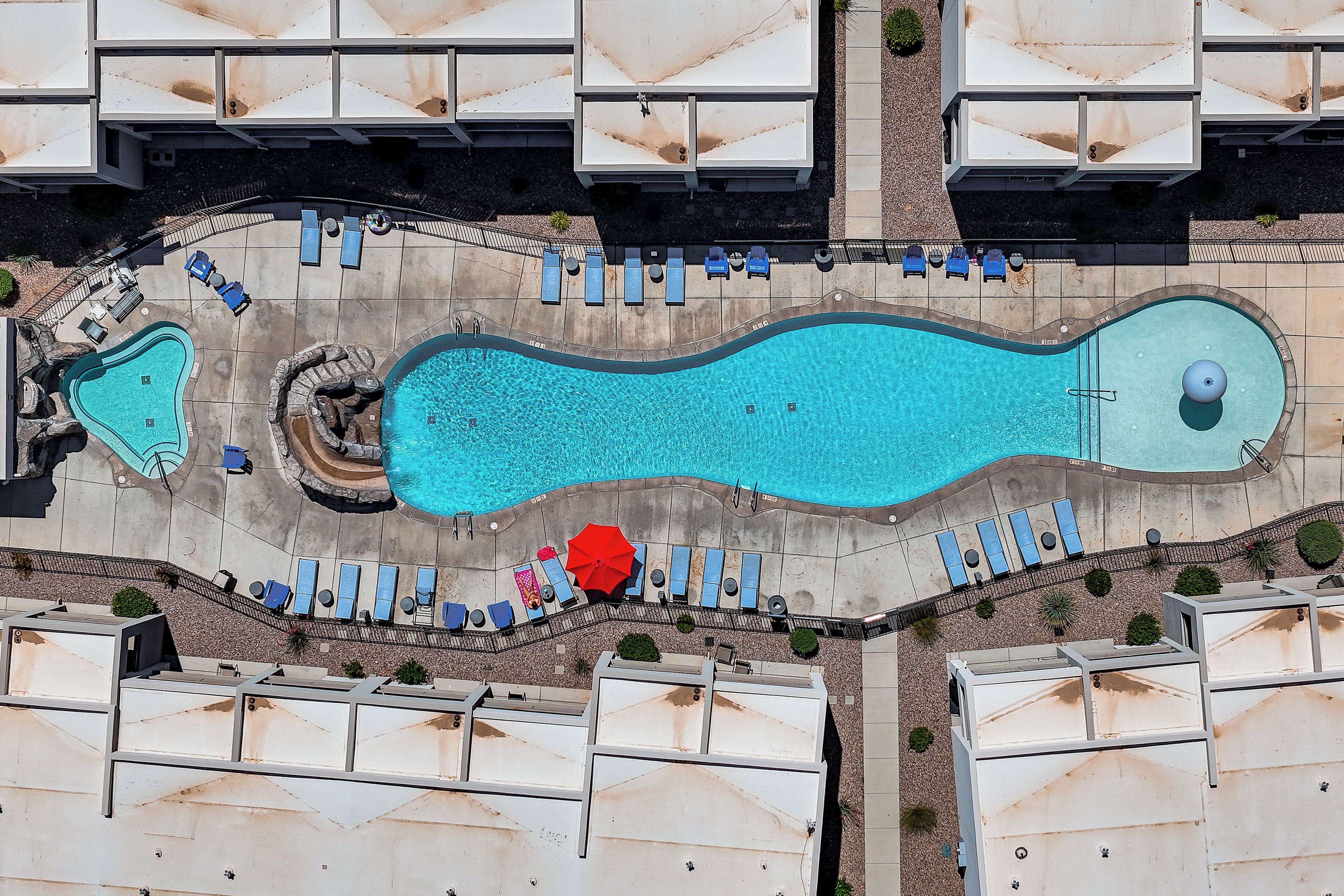 An aerial view of a resort with a large swimming pool, multiple lounge areas with umbrellas, and several guest rooms.
