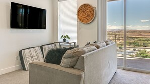 Un moderno salón con un sofá, un televisor de pantalla plana, una planta en maceta y vistas de un paisaje a través de amplios ventanales.