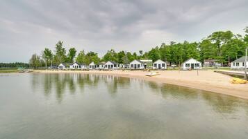 A row of white cabins with a sandy beach and a calm lake in the foreground.