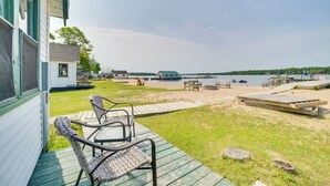 A deck with wicker chairs overlooking a lake, a dock, and a beach area with wooden benches and a picnic table.