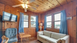 A cozy room with wooden walls, a ceiling fan, a flat-screen TV, a blue armchair, a beige sofa, and two windows with blue curtains. - Resort Cabin On Lake Huron! Pets Welcome (Cedarville)