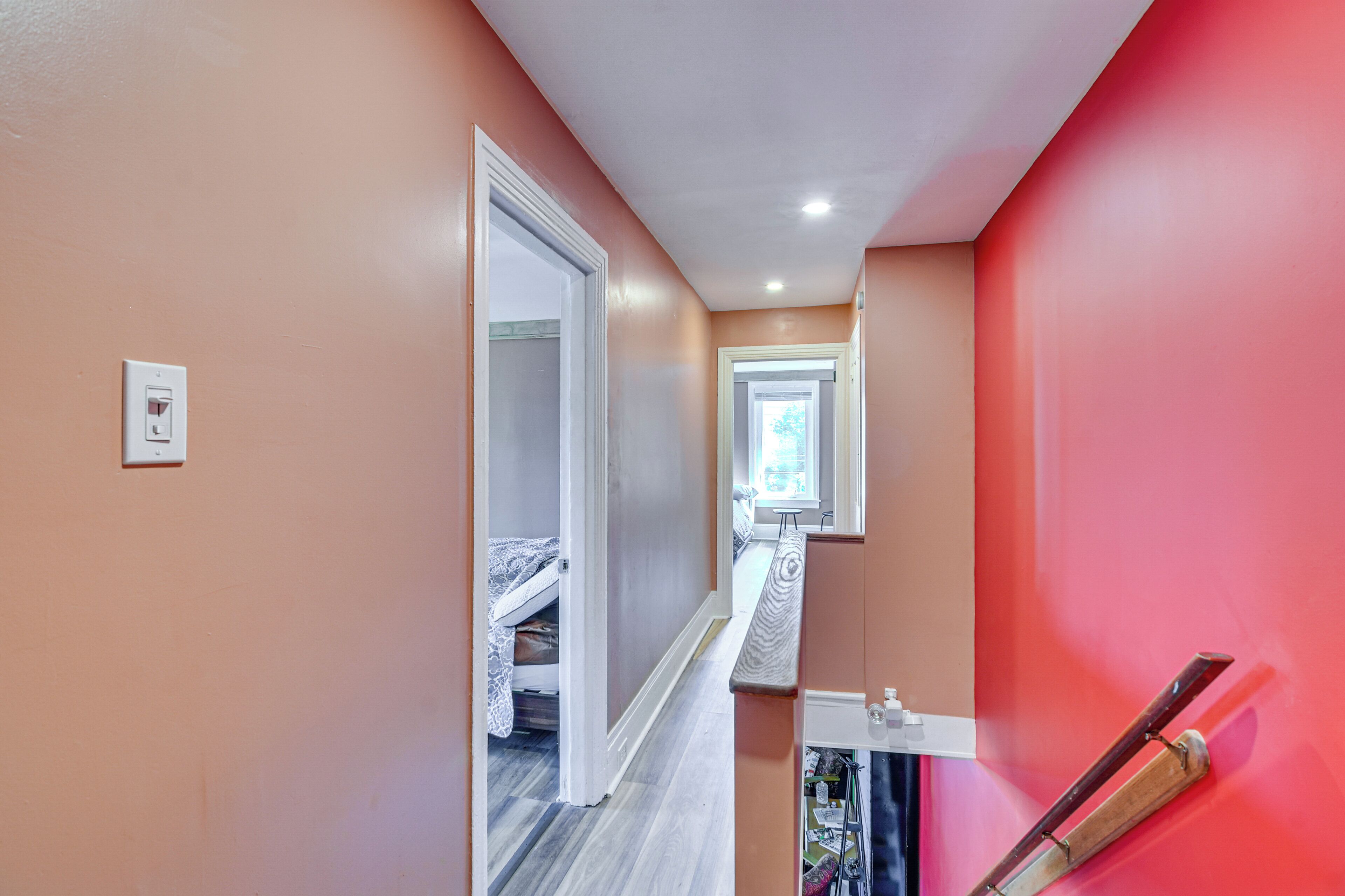 A narrow hallway with a red wall on one side and a white wall on the other. A door leads to a room with a bed and a window.