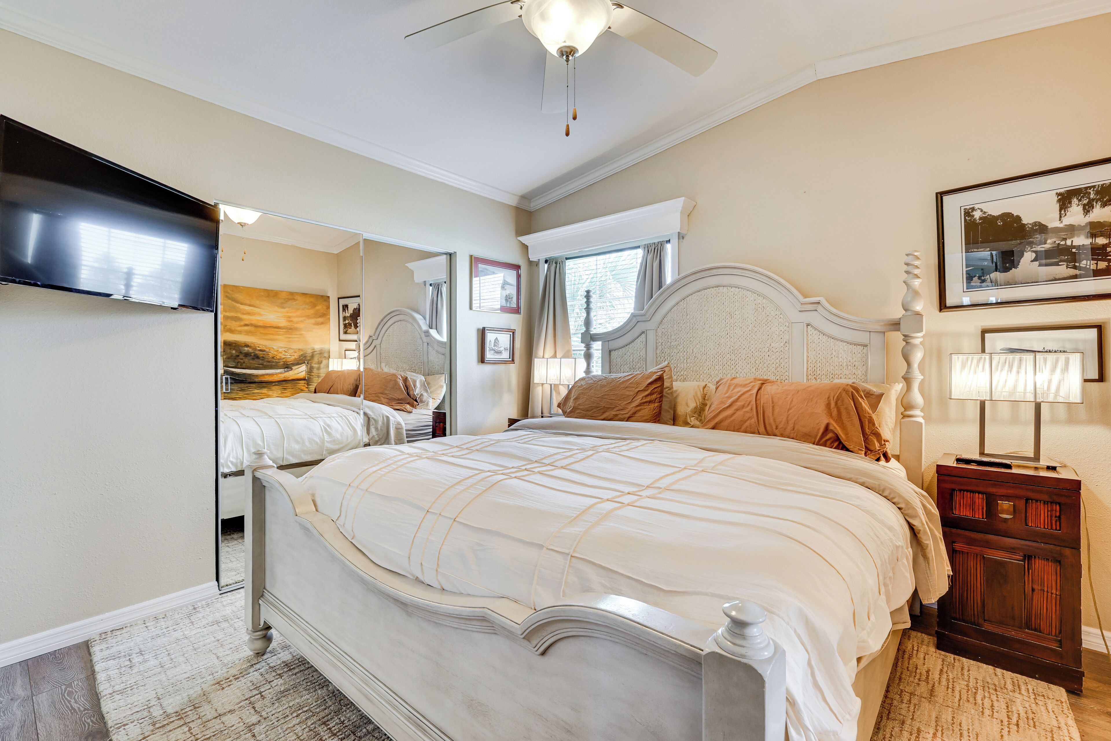 Une chambre avec un grand lit, un ventilateur de plafond, un téléviseur fixé au mur et une fenêtre avec des rideaux.