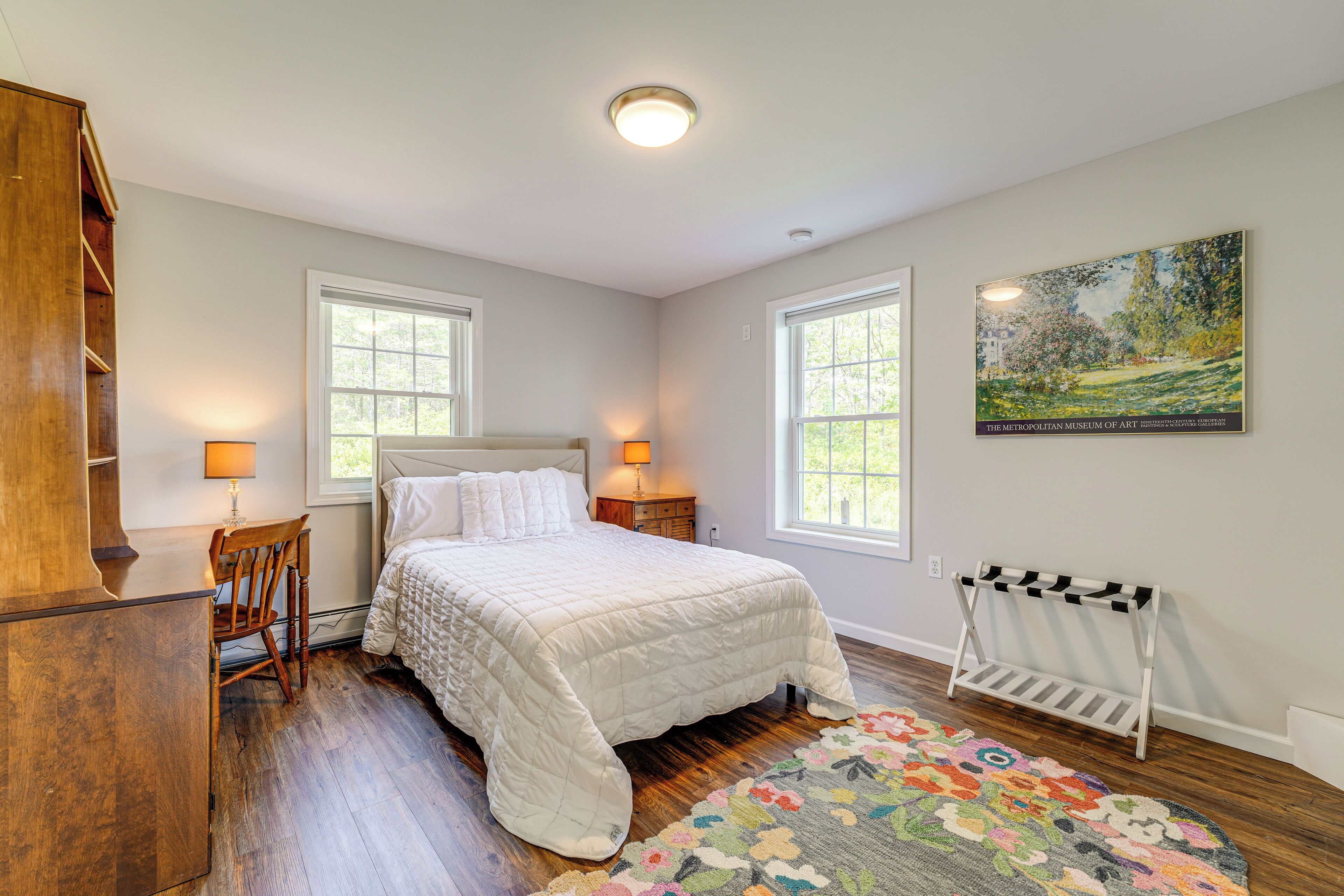 A bedroom with a bed, two bedside lamps, a wooden dresser, a chair, a radiator, and a painting on the wall.