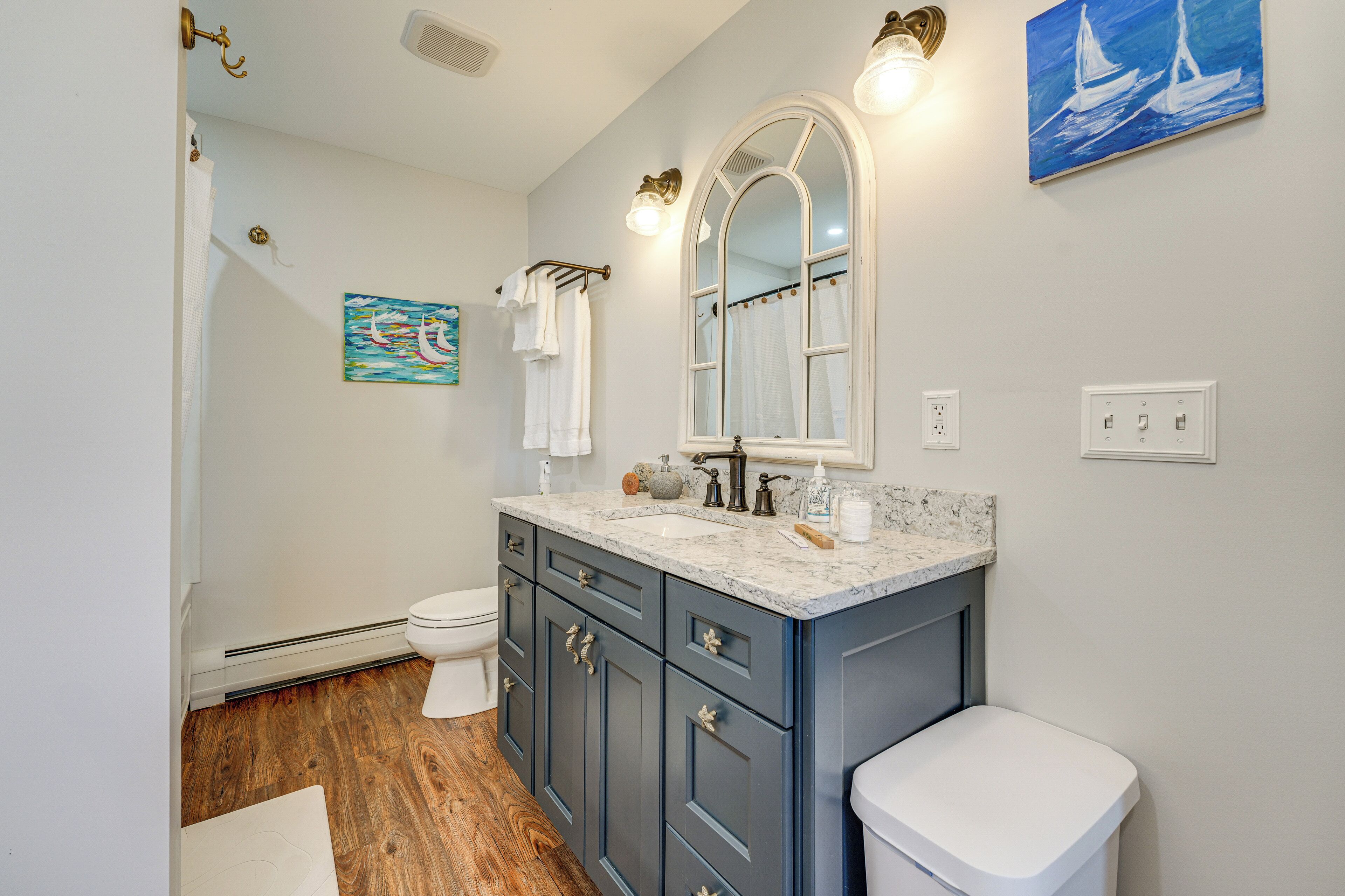 A bathroom with a marble sink, dark blue cabinets, a white toilet, and a painting of sailboats on the wall.
