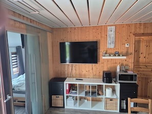 A room with a television mounted on the wall, a small kitchenette area with a microwave, and a bed with a window.
