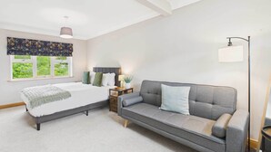 Un dormitorio con cama, sofá gris, mesita de noche, lámpara y ventana con vistas a zonas verdes.