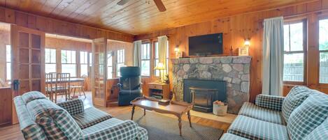 A cozy cabin interior with wooden walls, a stone fireplace, a ceiling fan, and plaid-patterned furniture.