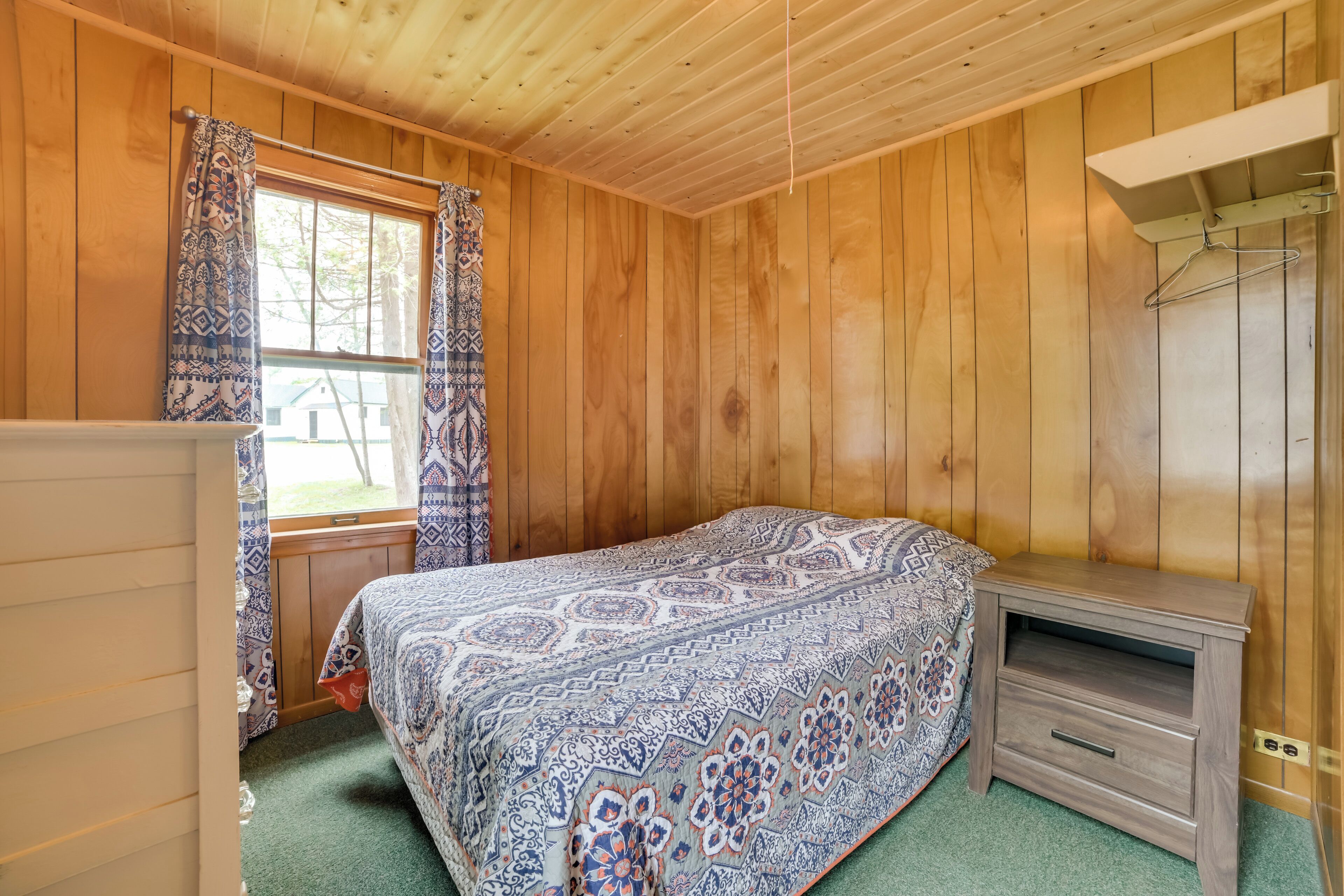 A wooden-paneled room with a bed, a dresser, a window with patterned curtains, and a small nightstand.