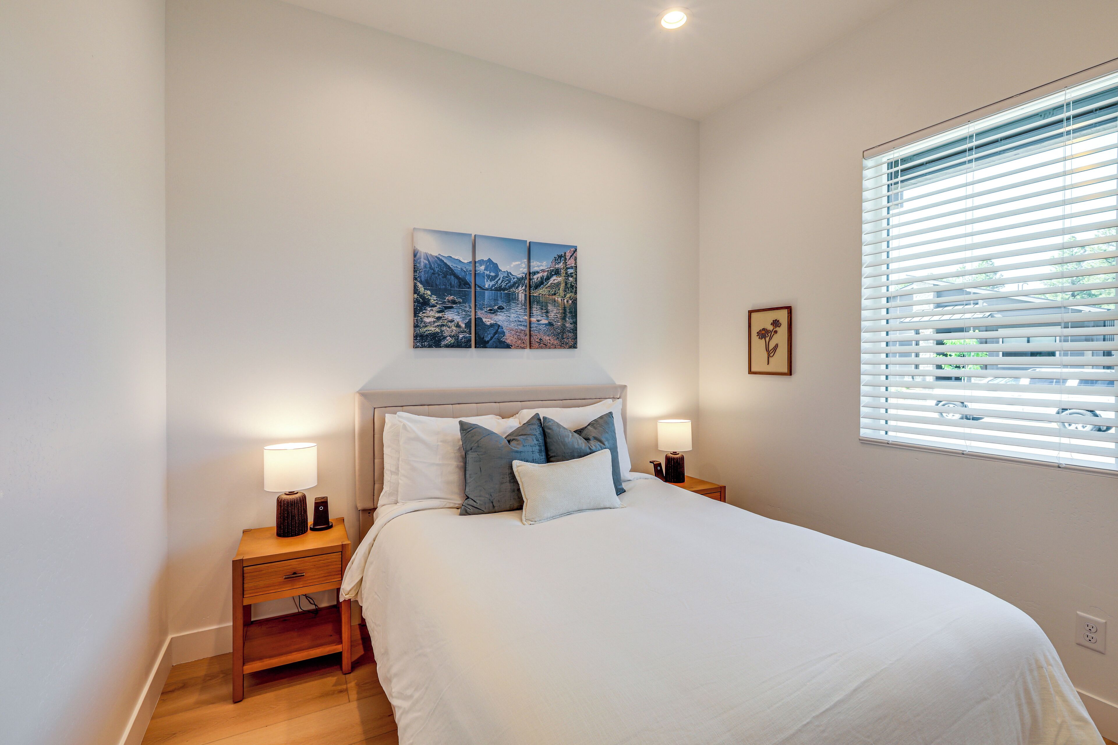 A bedroom with a bed, bedside tables, a nightstand, a window with blinds, and a wall-mounted picture.
