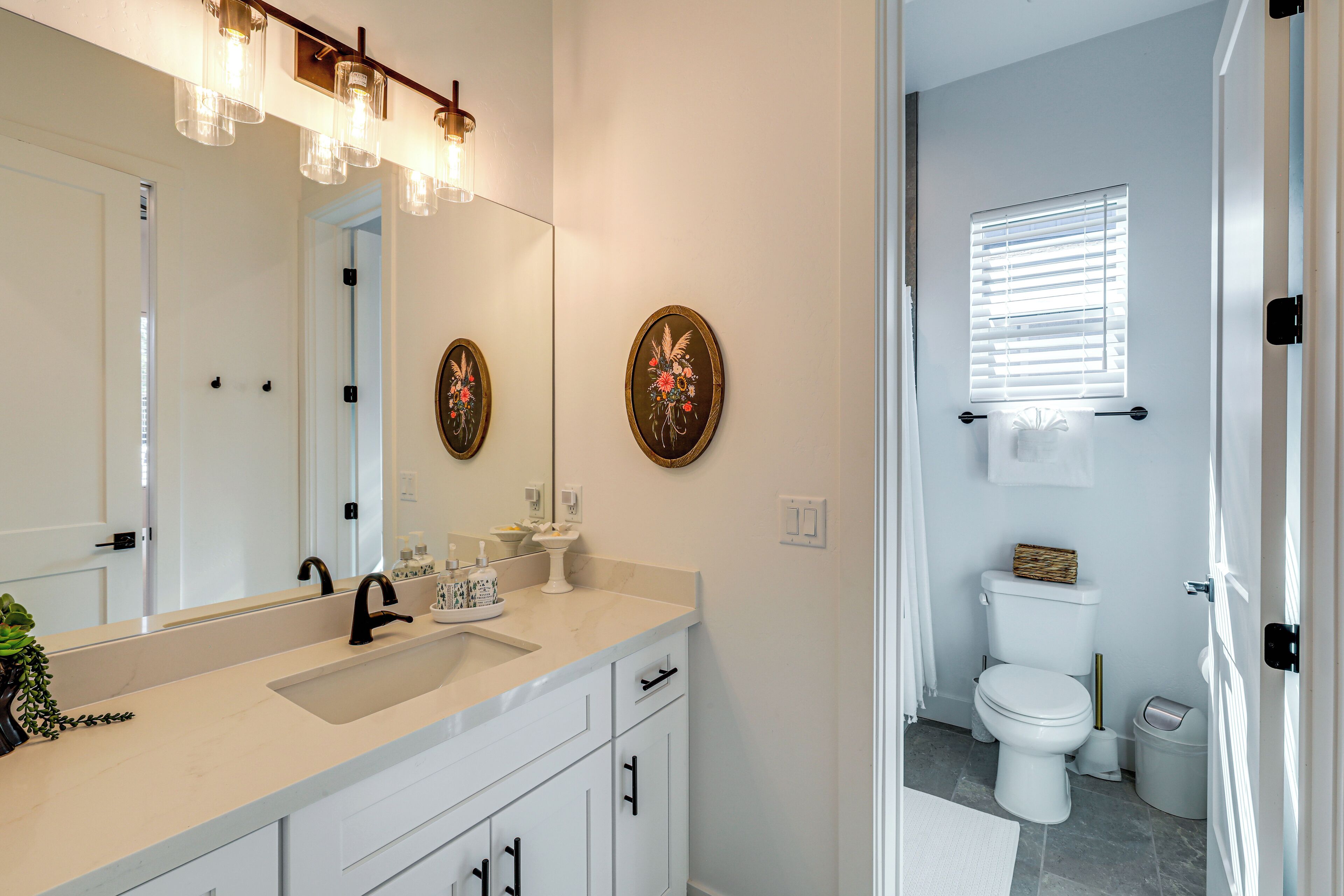 Un baño con lavamanos blanco, espejo y retrete. Hay platos decorativos en la pared y una ventana con persianas.