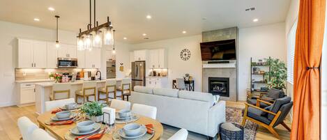 A modern kitchen with white cabinets, a dining area with a wooden table set for four, and a living room with a sofa and a fireplace.