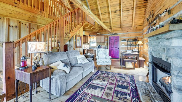 A cozy cabin interior with a wooden staircase, a fireplace, a dining area, and a living room furnished with a sofa and a coffee table.