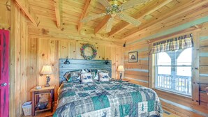 A wooden cabin bedroom with a bed, two bedside tables, a ceiling fan, a window with a view, and a door.
