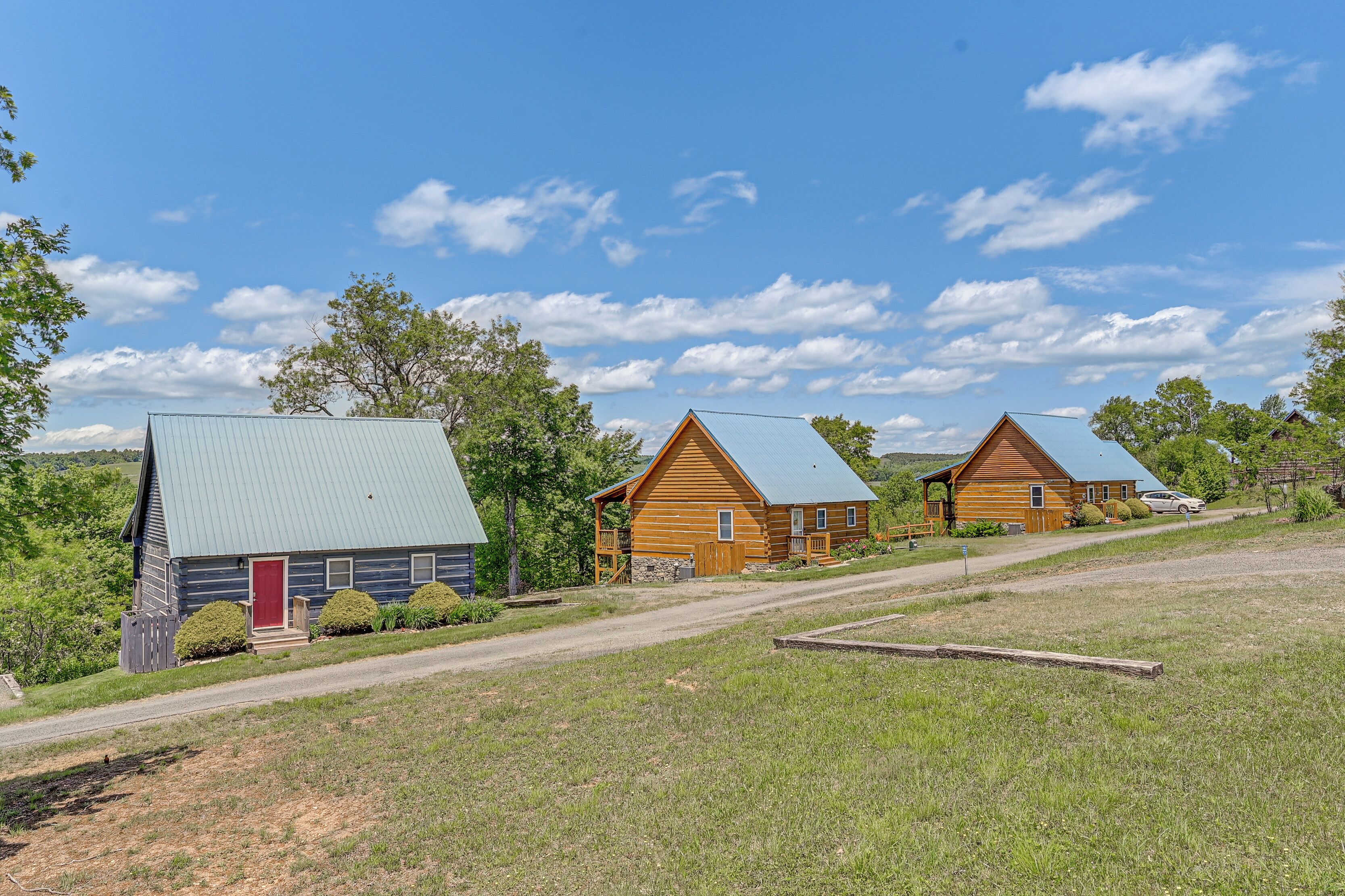 A row of log cabins with metal roofs, a dirt road, and a grassy field.