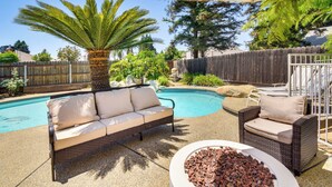 A backyard with a pool, a wicker sofa, a fire pit, and a wooden fence. - Clovis Family Home: Private Pool, Near Old Town! (Clovis)