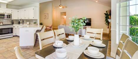 Una cocina moderna con armarios blancos, un comedor con mesa de madera preparada para cuatro personas y una sala de estar con sofá y televisor.